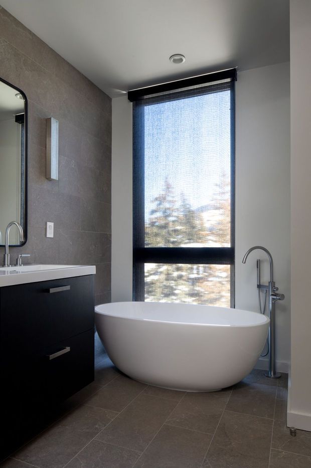 A contemporary bathroom featuring a freestanding white oval bathtub positioned in front of a large window with a black roller blind. The room has dark grey tiled walls and flooring, a black floating vanity with integrated basin, a rounded-edge mirror, and brushed metal tapware and fixtures.