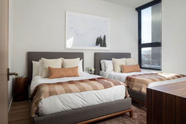 A warm, contemporary twin bedroom with two upholstered beds dressed in layered white, cream, and tan bedding. The room has a pale neutral wall, dark timber-look flooring, a large black-framed window, minimal framed artwork, a small side table, and a dark wood bedside or storage unit. Natural light creates a calm, refined atmosphere.
