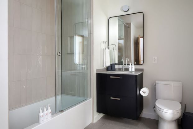 Bright, compact contemporary bathroom with a white tiled bathtub and glazed shower screen on the left, a dark floating vanity with a white basin and chrome tapware, a rounded rectangular black-framed mirror, and a white toilet. The room has a neutral palette with clean, practical finishes and ample natural light.