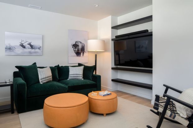 A contemporary living room with a deep green upholstered sofa, two round cognac-coloured ottomans, a black-and-white lounge chair, and a built-in entertainment wall with open shelving and a television. The room has white walls, light timber flooring, soft neutral carpeting, warm layered lighting, and restrained monochromatic artwork.