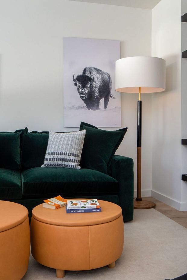 A cozy living room corner featuring a deep green velvet sofa with contrasting textured cushions, two rounded tan leather ottomans, a tall floor lamp, and a monochrome bison artwork. A neutral carpet and light walls are complemented by dark open shelving at the side.