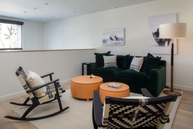 A bright, modern living room with a deep green sofa, two black-and-white patterned rocking chairs, and paired round tan ottoman-style coffee tables. The room has pale walls, light timber flooring, recessed ceiling lights, a large window, contemporary abstract artwork, and a tall floor lamp.