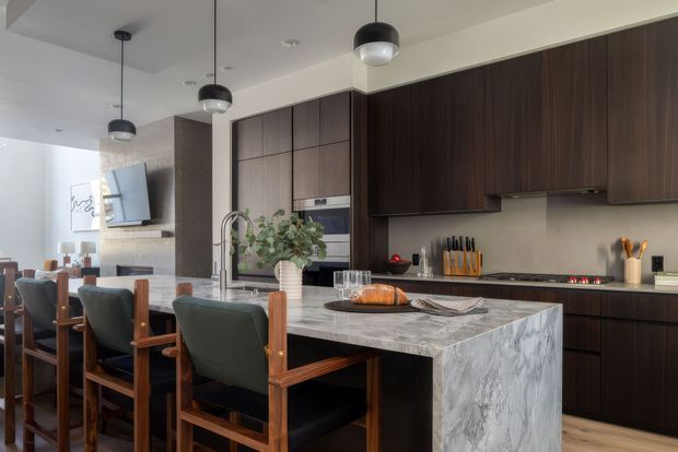 A refined open-plan kitchen and dining area with dark timber cabinetry, a large veined stone island, integrated appliances, pendant lighting, and timber-framed upholstered dining chairs. A living area with a television and fireplace wall is visible beyond the dining space.
