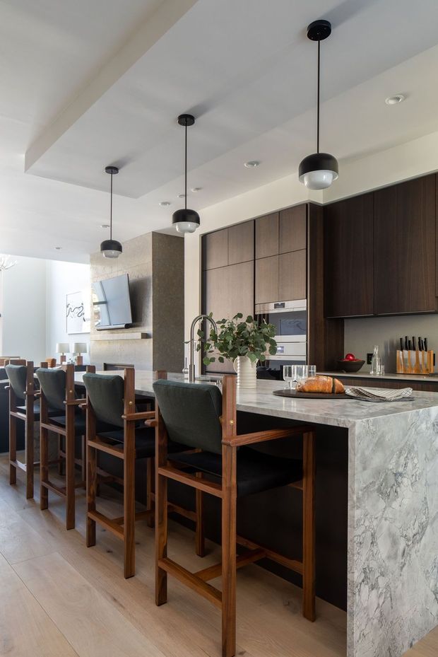 A refined open-plan kitchen and dining area with a large marble-look island, dark timber cabinetry, integrated appliances, and a row of black pendant lights. The island includes a sink and seating for several people on substantial timber-framed upholstered bar stools. A dining table and living area are visible beyond, creating a connected entertaining space. Light timber flooring and a pale ceiling balance the darker joinery and furniture.