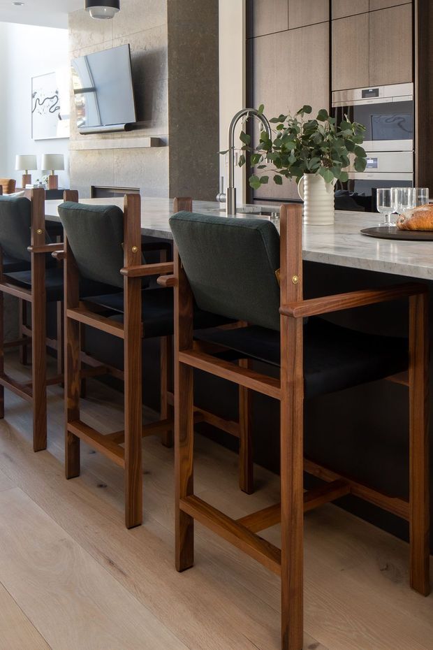 A refined open-plan kitchen and dining area with a long stone island, dark upholstered timber bar stools, integrated stainless-steel appliances, warm wood cabinetry, and a feature stone-clad wall. A dining table and living area are visible beyond, with soft natural light and restrained contemporary furnishings.
