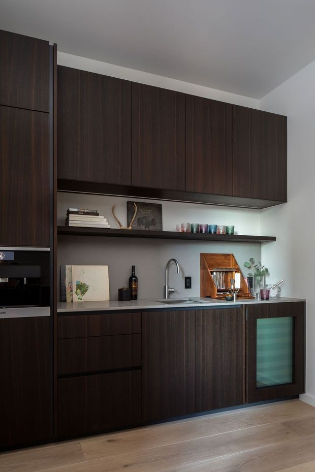 A compact contemporary kitchen or beverage station with floor-to-ceiling dark timber cabinetry, integrated appliances, open display shelving, a pale stone-look benchtop, undermount sink, and a glass-front beverage refrigerator. Light timber flooring and white walls provide contrast, while decorative glassware, books, and accessories add warmth.
