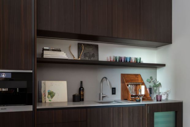 A refined contemporary kitchen with dark vertically grained timber cabinetry, a pale stone benchtop, integrated stainless-steel sink and brushed metal tapware. Open dark timber shelving displays books, glassware and decor, while under-cabinet lighting illuminates the grey backsplash. Built-in appliances and a small glass-fronted wine or drinks refrigerator complete the compact, highly tailored joinery.