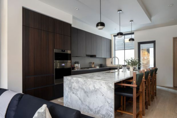 A contemporary open-plan kitchen and dining area viewed from the living space. The kitchen features dark timber-toned full-height cabinetry, integrated appliances, a large pale veined stone island with waterfall edge, dark pendant lights, and a timber dining table with upholstered chairs beside a glazed exterior door. Light timber flooring and white walls create a balanced, refined interior.