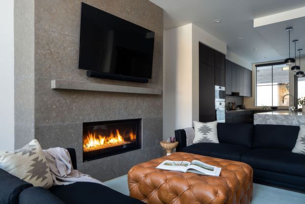 A contemporary open-plan living room with a dark sectional sofa, matching armchair, tufted leather ottoman, and a wall-mounted television above a built-in linear fireplace. The fireplace wall is finished in textured stone, while a dark contemporary kitchen with a stone island and pendant lights is visible beyond.