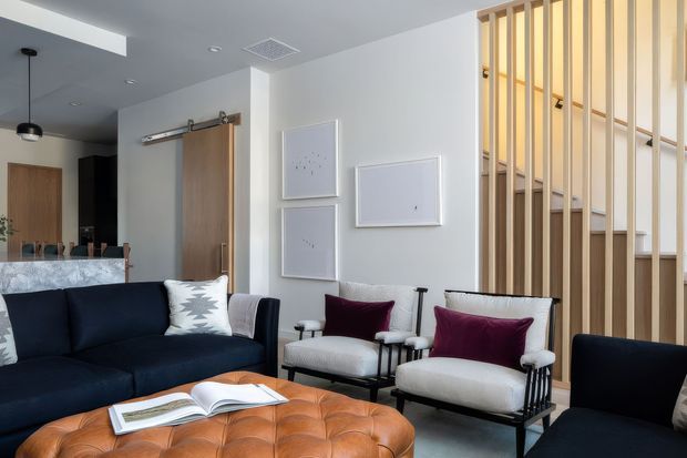 Contemporary open-plan living room with a deep navy sofa, two light upholstered black-framed armchairs, burgundy accent cushions, and a large cognac leather tufted ottoman. A timber barn-style sliding door, white walls, recessed lighting, and a vertical timber slat stair balustrade create a clean, modern interior. The kitchen and dining area are visible in the background.