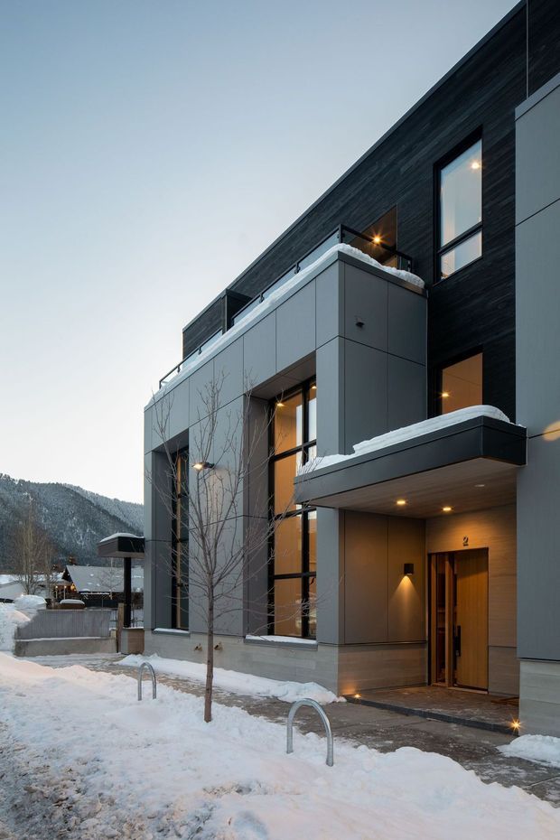 Exterior architectural photograph of a contemporary multi-storey residential building in a snowy mountainous setting. The facade combines dark horizontal cladding with smooth grey panel sections, tall black-framed windows, projecting balconies, recessed warm lighting, and a timber-toned entry door. Snow-covered ground, neighboring homes, and a forested mountain are visible in the background.
