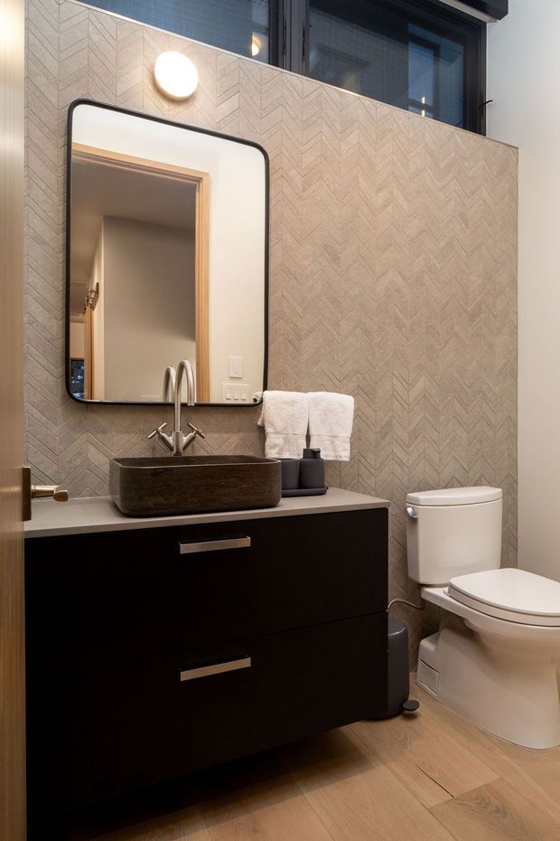 Compact contemporary bathroom with a dark floating vanity, vessel basin, chrome wall-mounted tapware, rounded rectangular mirror, white toilet, and warm timber-look flooring. The walls feature a beige-grey chevron-patterned tile finish, with a wall sconce and a small upper window providing light.