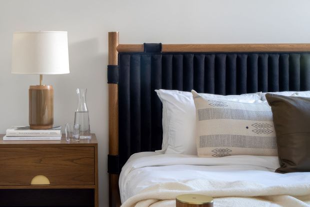 Close-up of a refined bedroom corner featuring a bed with a tall vertically channelled dark upholstered headboard framed in warm timber, crisp white bedding, patterned neutral cushions, and a mid-century-inspired timber bedside table with a ceramic lamp, books, and glassware. The styling is calm and understated with warm natural wood and deep navy-black accents.
