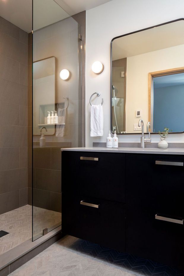 Contemporary bathroom with a walk-in glass shower, large rounded-corner mirror, wall-mounted circular sconces, black vanity cabinetry with a pale benchtop, chrome fixtures, and light patterned floor and wall tiles. A blue-painted adjoining room is visible through the doorway reflected in the mirror.
