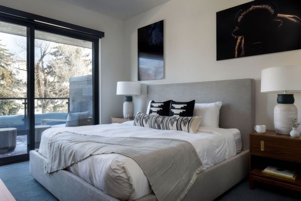 A stylish contemporary bedroom with a large upholstered bed, layered white and grey bedding, patterned accent cushions, and matching bedside tables with ceramic lamps. Black-framed floor-to-ceiling glazing opens to an outdoor balcony or terrace, while monochrome artwork adds contrast to the neutral scheme.