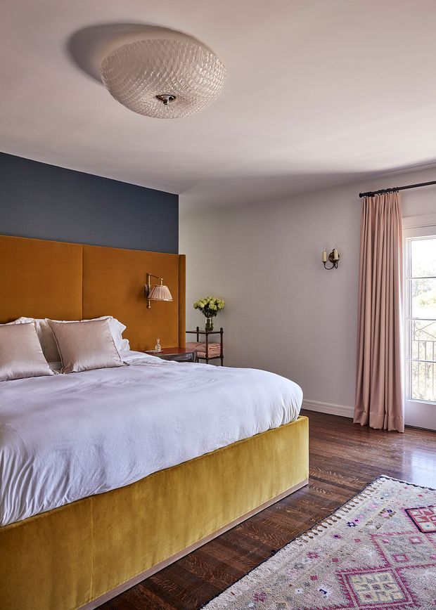 A refined bedroom with a large upholstered mustard-gold bed and matching headboard, pale lavender-grey bedding, a deep blue accent wall, pink floor-length curtains, dark timber flooring, and a patterned area rug. Decorative wall and ceiling lighting, a small bedside table, and a glazed balcony door complete the warmly styled room.