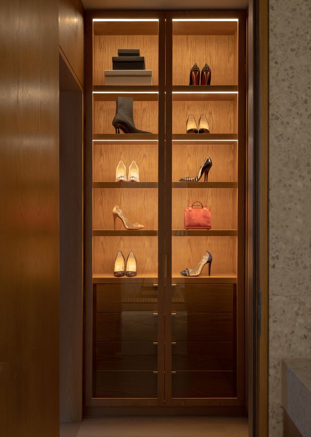 A tall built-in display wardrobe with glass doors, warm timber veneer shelving, integrated linear lighting, and dark timber drawer fronts. The shelves display designer shoes, boots, handbags, and accessories. The cabinetry is set within a refined interior with a textured stone or patterned wall visible at the right edge.