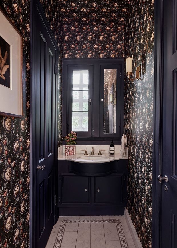 A compact, dramatically styled powder room with dark navy panelled doors and vanity cabinetry, ornate dark floral wallpaper covering the walls and ceiling, a pale stone or marble countertop with integrated basin, brass tapware and wall sconce, and a patterned light tile floor. A window and narrow mirror sit above the vanity.