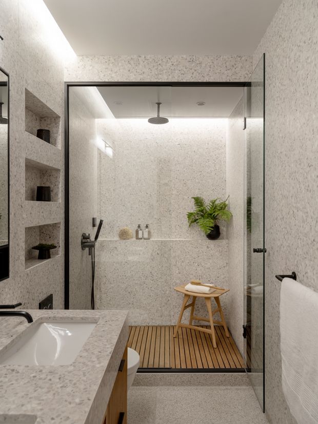 Contemporary bathroom featuring a fully tiled walk-in shower enclosed by clear glass, integrated wall niches, a rainfall showerhead, wall-mounted controls, a floating vanity with a pale terrazzo-like benchtop and undermount basin, and a timber slatted shower floor. Recessed and concealed lighting creates a soft, refined atmosphere, with a potted fern adding a natural accent.