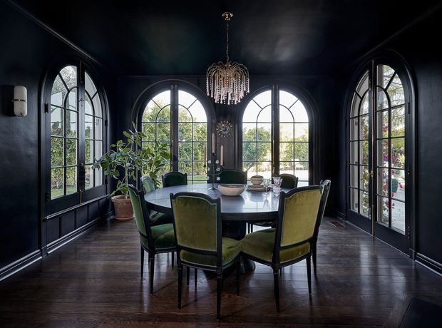 A dramatic, moody dining room with a dark painted interior, three large arched multi-pane windows and glazed doors, and dark timber flooring. A round dining table is surrounded by upholstered green dining chairs, with a statement crystal chandelier overhead and potted greenery near the windows.