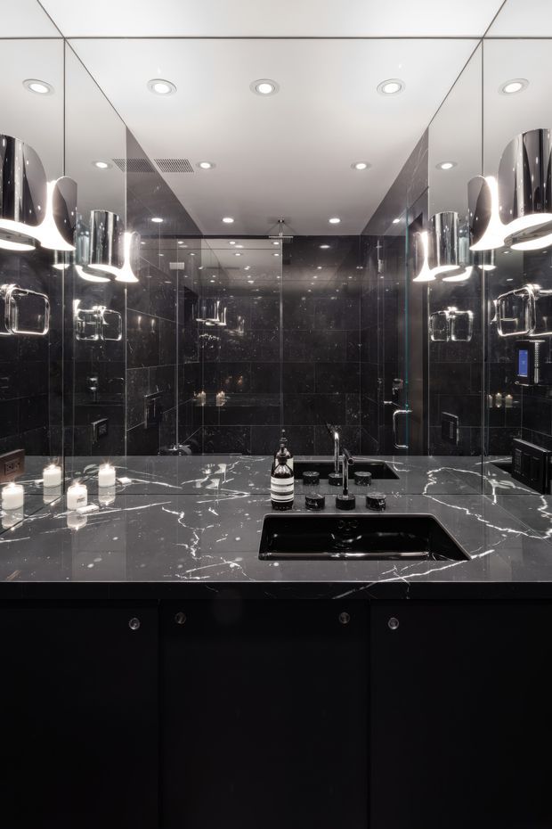 A dramatic, luxurious bathroom vanity viewed straight on. The space features a large mirrored wall, glossy black stone wall tiles, a dark marble-look benchtop with prominent white veining, an integrated black basin, matte black cabinetry, chrome fixtures, and sculptural pendant lights. Recessed ceiling lights provide additional illumination, creating a high-contrast, polished interior.