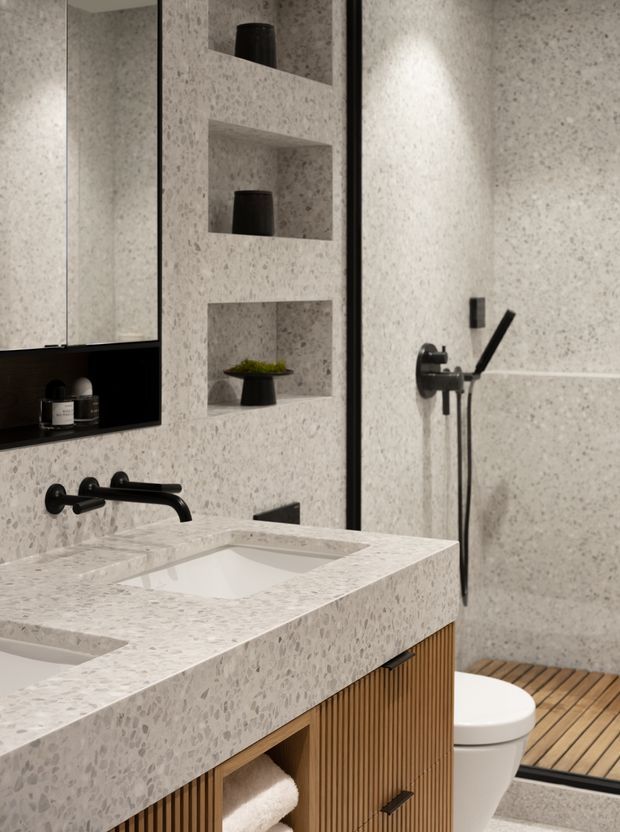 Contemporary bathroom with a terrazzo-look tiled wall finish and matching thick vanity benchtop, integrated white basins, matte black wall-mounted tapware and shower fittings, recessed display niches, and a warm timber fluted vanity. A frameless shower enclosure with a timber-look slatted floor and a partially visible toilet complete the space.