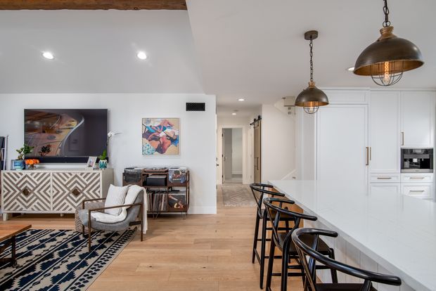 Open-plan contemporary interior combining a living room and kitchen. The space features white walls and cabinetry, light timber flooring, a large white kitchen island with black bar stools, dark metal pendant lights, a patterned rug, a geometric console, exposed timber ceiling beam, and artwork.