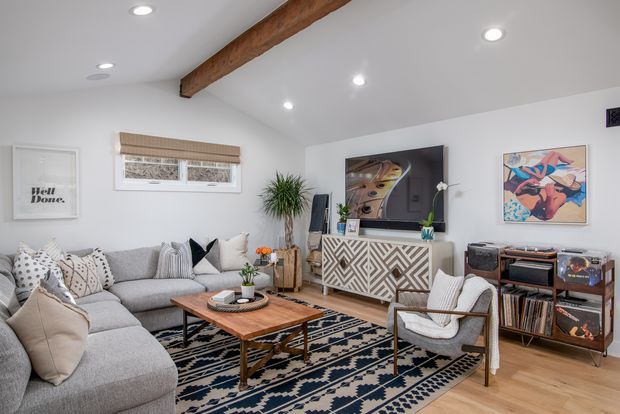 Bright, inviting living room with a large L-shaped grey sectional sofa, patterned area rug, wooden coffee table, woven window blind, media console, television, record player and vinyl storage. White walls and ceiling are complemented by a prominent exposed timber ceiling beam, indoor plants, artwork and layered neutral textiles.
