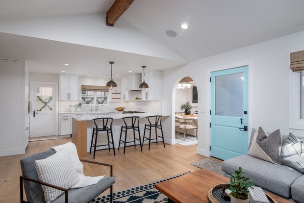 Bright open-plan living area with a white kitchen, timber island and three bar stools, adjoining a dining nook and a seating area with a grey sofa, armchair and timber coffee table. The room has light timber flooring, white cabinetry and walls, exposed timber ceiling beam, pendant lights, an arched opening, and a distinctive turquoise-blue entry door.