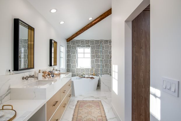 Bright, spacious bathroom with a freestanding white bathtub, double vanity, marble-look surfaces, warm wood cabinetry, and a bold black-and-white geometric feature wall. A timber ceiling beam, recessed lighting, mirrors, and a patterned runner add contrast and character.