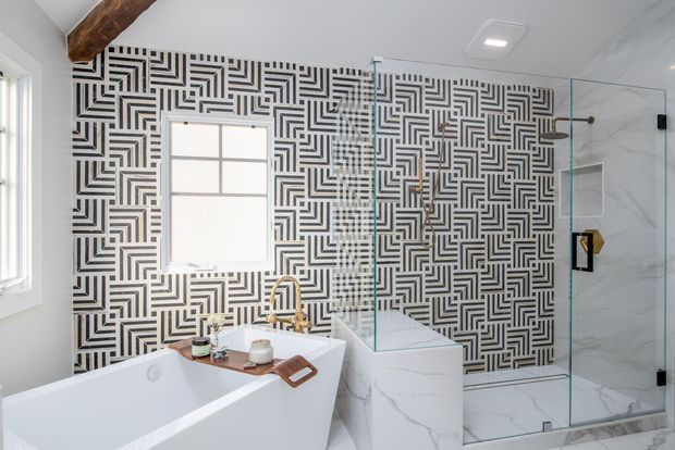 Bright contemporary bathroom featuring a freestanding white bathtub, glass-enclosed shower, marble-look surfaces, and a bold black-and-white geometric patterned feature wall. A timber ceiling beam adds warmth and contrast.