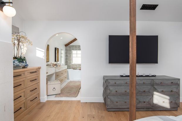 Bright contemporary bedroom interior with warm timber flooring, a large rustic timber dresser, wall-mounted television, and a vertical exposed timber post. An arched opening leads to a bathroom or dressing area with a double vanity, patterned feature wall, and freestanding bathtub. The room includes a ceiling-mounted light fixture and abundant natural light.