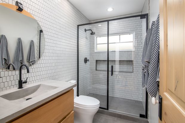 Bright, compact bathroom with white small-format tiled walls, a grey tiled shower enclosure with black-framed glass, a floating timber vanity with integrated basin, black tapware, recessed shower niches, and a timber door. A wall mirror and striped towels add contrast.