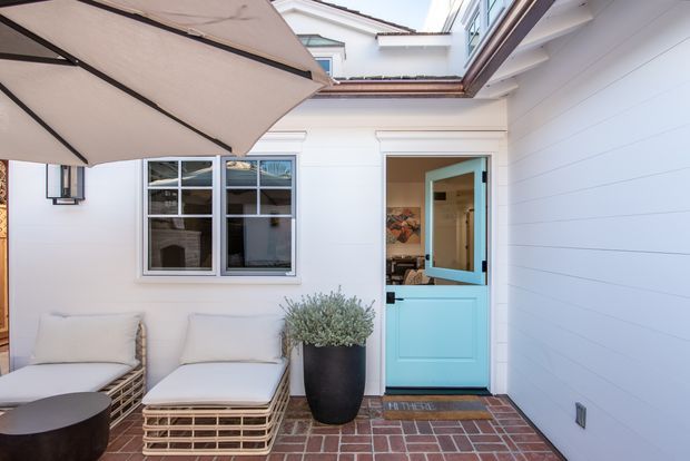 Bright residential courtyard or entry area with white horizontal weatherboard walls, a vivid turquoise panelled door, multi-pane window, red brick paving, wicker lounge chairs, a large outdoor umbrella, and a potted plant. The space has a relaxed coastal character and appears to be viewed from within the courtyard toward the entrance.