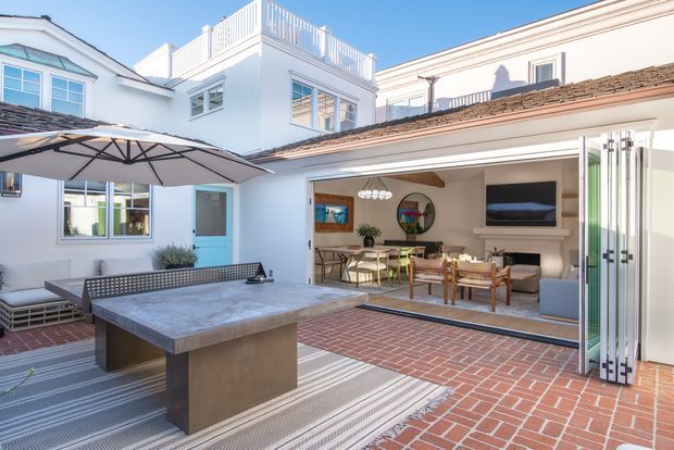 A bright, spacious residential courtyard centered on a built-in outdoor table-tennis table beneath a large umbrella. White rendered multi-level buildings surround the courtyard, with red brick paving, an outdoor dining area, and a large open glazed folding-door connection to an interior living and dining space. The interior includes a fireplace, television, dining table, and exposed timber ceiling beam.