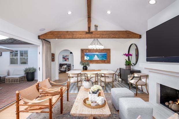 Bright open-plan living and dining room with white walls, exposed rustic timber beam and central post, a large wall-mounted television above a white fireplace, dining table with upholstered chairs, lounge seating, leather sling chairs, and a pale timber coffee table. An arched opening leads toward an adjacent seating area, while glazed doors open to a brick-paved outdoor space. The room combines relaxed contemporary furnishings with rustic structural detailing.