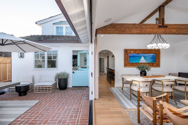 A bright indoor-outdoor dining area with large folding or sliding glass doors opening onto a brick-paved courtyard. The interior has a timber floor, white walls, exposed rustic timber beams, a long dining table with sculptural pendant lighting, and an arched opening leading toward the kitchen. Outside, a white-clad cottage-style facade features a light blue door, multi-pane windows, outdoor seating, and a patio umbrella.