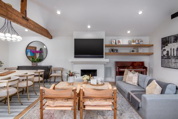 Bright open-plan living and dining room with a white fireplace wall, wall-mounted television, exposed rustic timber ceiling beams, floating timber display shelves, grey sectional sofa, timber dining table and chairs, and layered neutral furnishings. A piano and framed wall art are visible on the right.