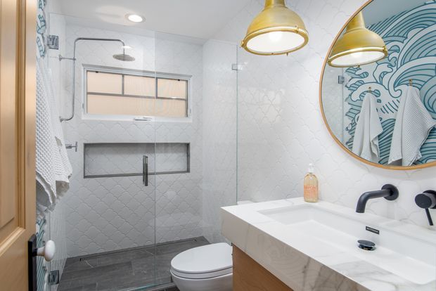 Bright contemporary bathroom with a glass-enclosed walk-in shower, wall niche, toilet, double vanity, marble-look countertop, black fixtures, white patterned wall tiles, and large round mirror. Brass pendant lights and a blue wave-patterned mirror backdrop add decorative accents.