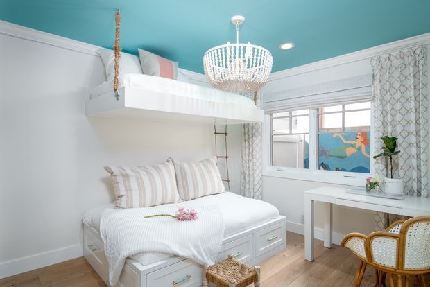 Bright, compact bedroom with a built-in lower bed and elevated loft bed, white built-in drawers, a rope ladder, patterned curtains, and a small white desk by the window. The turquoise ceiling, crystal chandelier, striped bedding, and rattan chair create a playful coastal-inspired interior.