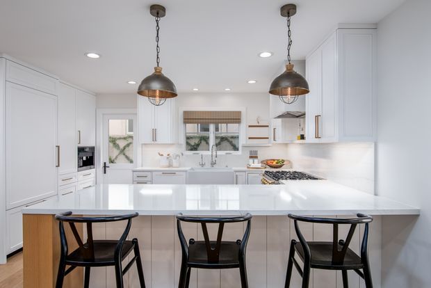 Bright, spacious white kitchen with extensive shaker-style cabinetry, a large central island with integrated seating, white benchtops, stainless-steel appliances, and two dark industrial pendant lights. Black timber bar stools provide contrast against the predominantly white and light-wood palette.