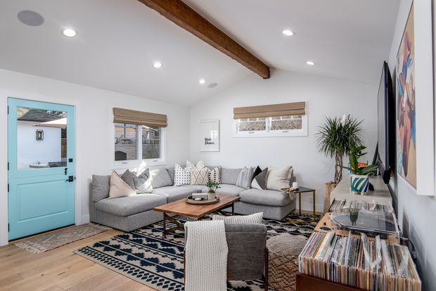Bright, cozy living room with a large grey sectional sofa, patterned black-and-white rug, timber coffee table, armchair, record storage, indoor plant, and colorful wall art. The room has white walls, a white ceiling with recessed lighting, exposed timber ceiling beam, light timber flooring, and a distinctive turquoise exterior door opening to a small outdoor area.