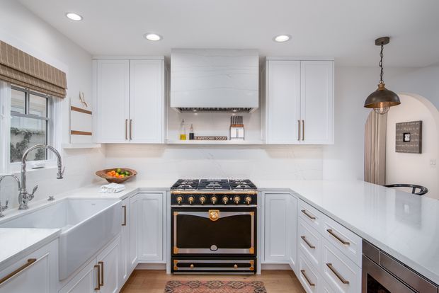 Bright, refined kitchen with white raised-panel cabinetry, a large marble-look range hood, professional range cooker, pale stone-look benchtops and splashback, farmhouse sink, brass hardware, and a warm metallic pendant. An arched opening leads to an adjacent area, while a patterned runner adds colour to the timber-toned floor.