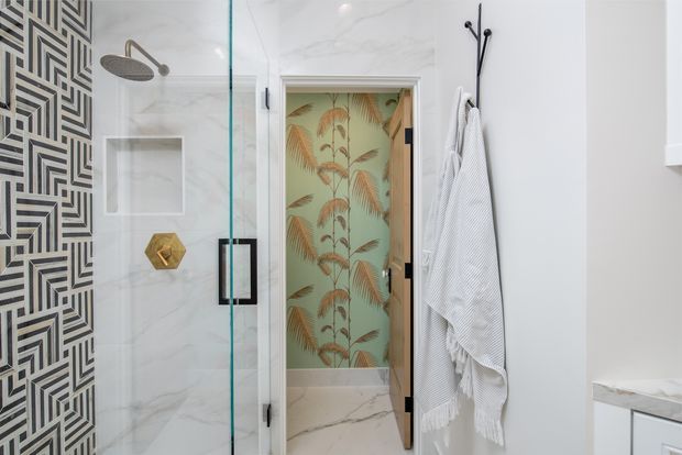 A refined contemporary bathroom with a frameless glass shower enclosure, geometric black-and-white patterned tile feature wall, marble-look wall and floor surfaces, and brushed metal shower fittings. A doorway opens to a leafy botanical wallpapered space, while white towels hang from black wall hooks.