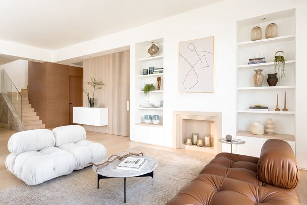 Bright, refined living room with a sculptural white modular sofa and brown leather sectional arranged around a round stone coffee table. Built-in white shelving displays ceramics and books beside a simple fireplace with a pale surround, while timber wall panelling and a slatted feature wall frame the entry and stairway. The room has a light neutral palette, ample natural light, and a contemporary high-end residential feel.
