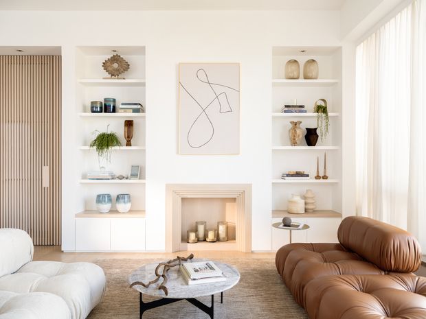 Bright, refined living room with built-in white shelving flanking a decorative fireplace, sculptural wall art, curated ceramics and books, a large brown leather sofa, white upholstered lounge seating, and a round stone coffee table. Full-height sheer curtains bring in abundant daylight, while a warm neutral rug and restrained contemporary decor create a calm, polished interior.