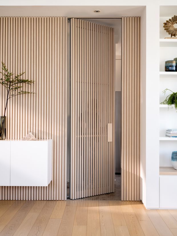 A refined interior entry or hallway featuring a full-height vertical timber-slat feature wall and matching pivot door. A slim white floating cabinet, indoor tree, recessed ceiling light, and white open shelving with decorative objects frame the composition. Pale timber flooring and warm neutral tones create a clean, contemporary and softly textured space.