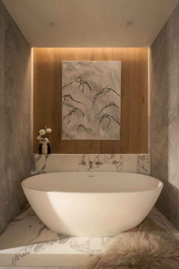 A luxurious, warm-toned bathroom featuring a freestanding sculptural bathtub set on a raised marble platform. Veined marble covers the side walls and floor, while a timber feature wall with concealed warm LED lighting frames a minimalist mountain artwork. Brushed metal wall-mounted tapware, recessed ceiling lights, and a small decorative arrangement complete the refined, spa-like setting.