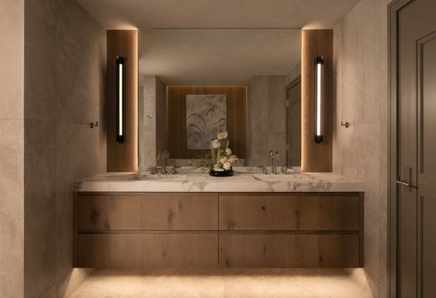 A refined, warm-toned bathroom vanity viewed straight on. The room features large beige stone-look wall and floor surfaces, a floating dark timber double vanity with a pale veined stone benchtop, two integrated basins and taps, a large central mirror, and tall vertical wall sconces framed by timber-toned panels. A framed artwork and floral arrangement sit on the vanity, while a dark paneled door is visible at right.