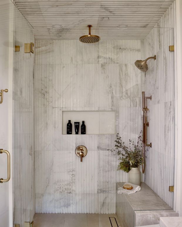 A refined walk-in shower with vertically fluted pale marble or stone wall surfaces, a built-in recessed shelf, brushed brass rain shower and handheld shower fittings, a built-in tiled bench, and a linear floor drain. The space is bright, neutral, and softly luxurious, with a floral arrangement and bath accessories on the bench.